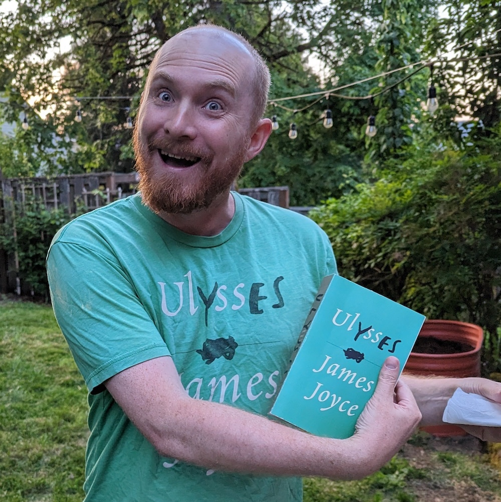 A bearded, bald man with a wide-eyed, wild look holding a green copy of Ulysses by James Joyce which matches his t-shirt.