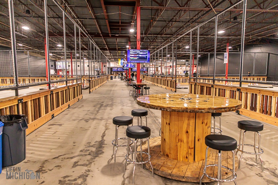 Interior view of the Fowling Warehouse with a wooden high-top table and stools in the foreground, fenced in fowling lanes on either side, and a warm-lit industrial ceiling.